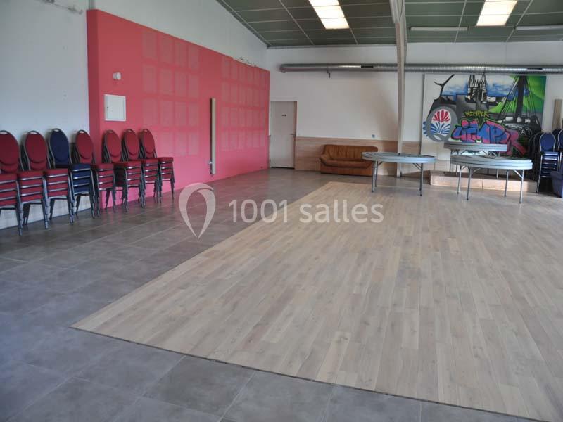 Salle polyvalente vide avec sol en carrelage et parquet, chaises empilées, tables pliées et fresque murale colorée.