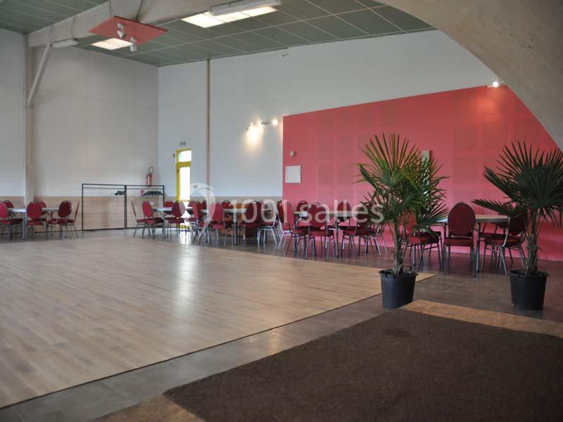 Salle lumineuse avec parquet, chaises rouges alignées, tables noires et plantes en pot devant un mur rose.