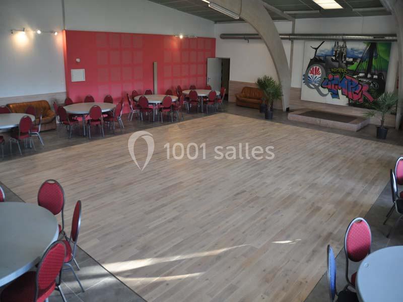 Salle spacieuse avec parquet clair, tables et chaises rouges disposées autour, scène en fond avec fresque colorée.