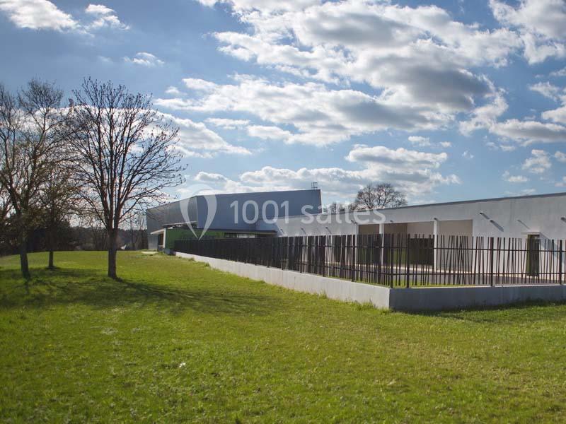 Bâtiment moderne entouré d'une pelouse verte, avec une clôture métallique et un ciel partiellement nuageux.