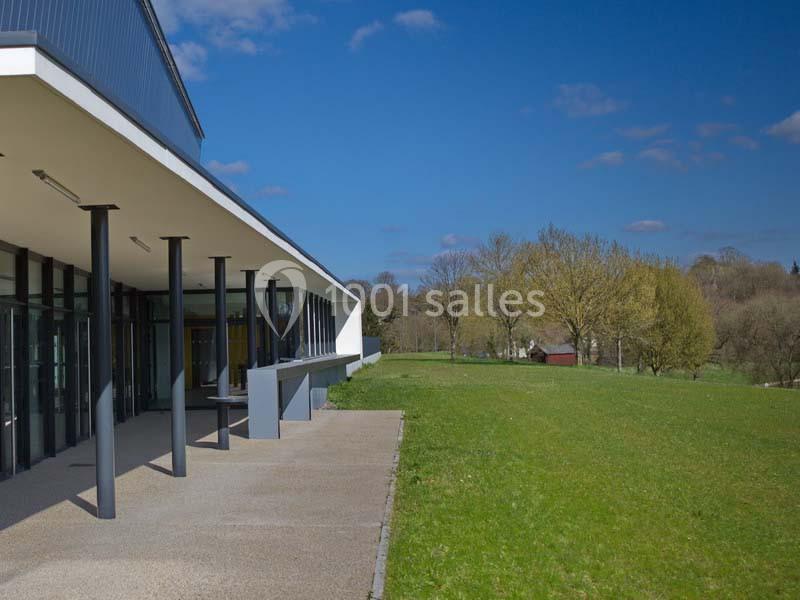 Bâtiment moderne avec une terrasse couverte, donnant sur une pelouse et des arbres sous un ciel bleu.