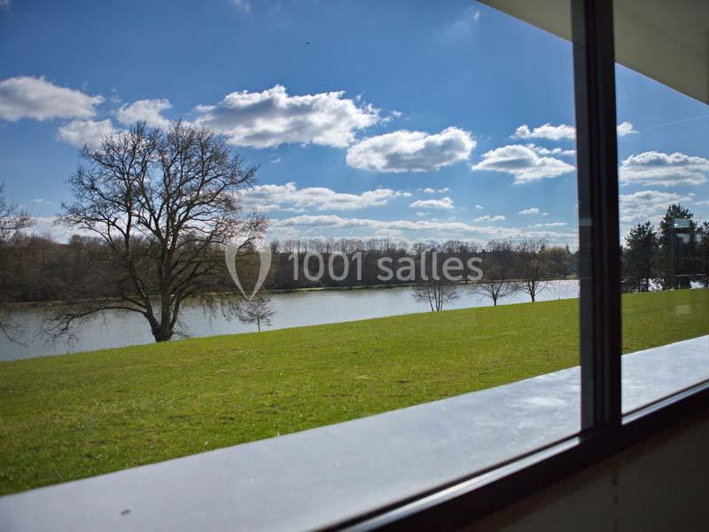 Vue depuis une fenêtre sur un parc avec une pelouse, des arbres, un lac et un ciel bleu parsemé de nuages.