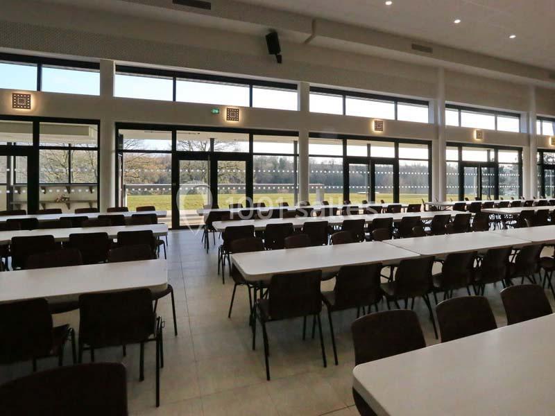 Salle spacieuse avec de grandes baies vitrées, des tables alignées et des chaises, donnant sur un espace extérieur verdoyant.