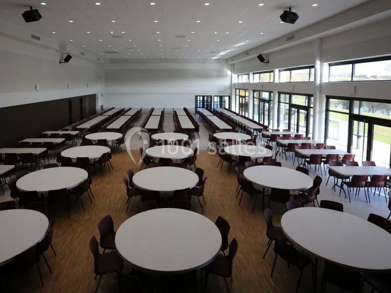 Salle spacieuse avec des tables rondes blanches et des chaises disposées en rangées, éclairée par de grandes baies vitrées.