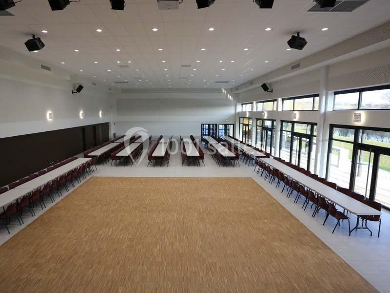 Salle spacieuse avec tables et chaises alignées, parquet central et grandes baies vitrées laissant entrer la lumière…