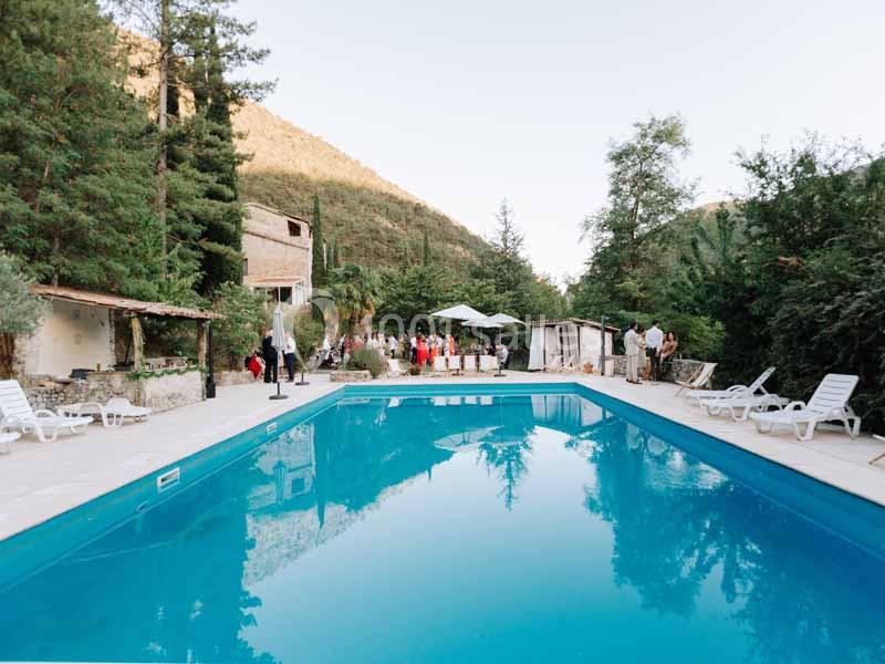 Piscine extérieure entourée de chaises longues, arbres et bâtiments, avec des personnes en arrière-plan.