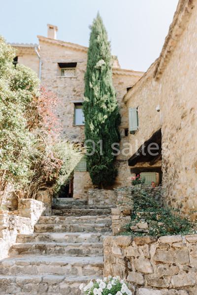 Escalier en pierre bordé de végétation menant à des bâtiments en pierre avec volets verts et un grand cyprès.