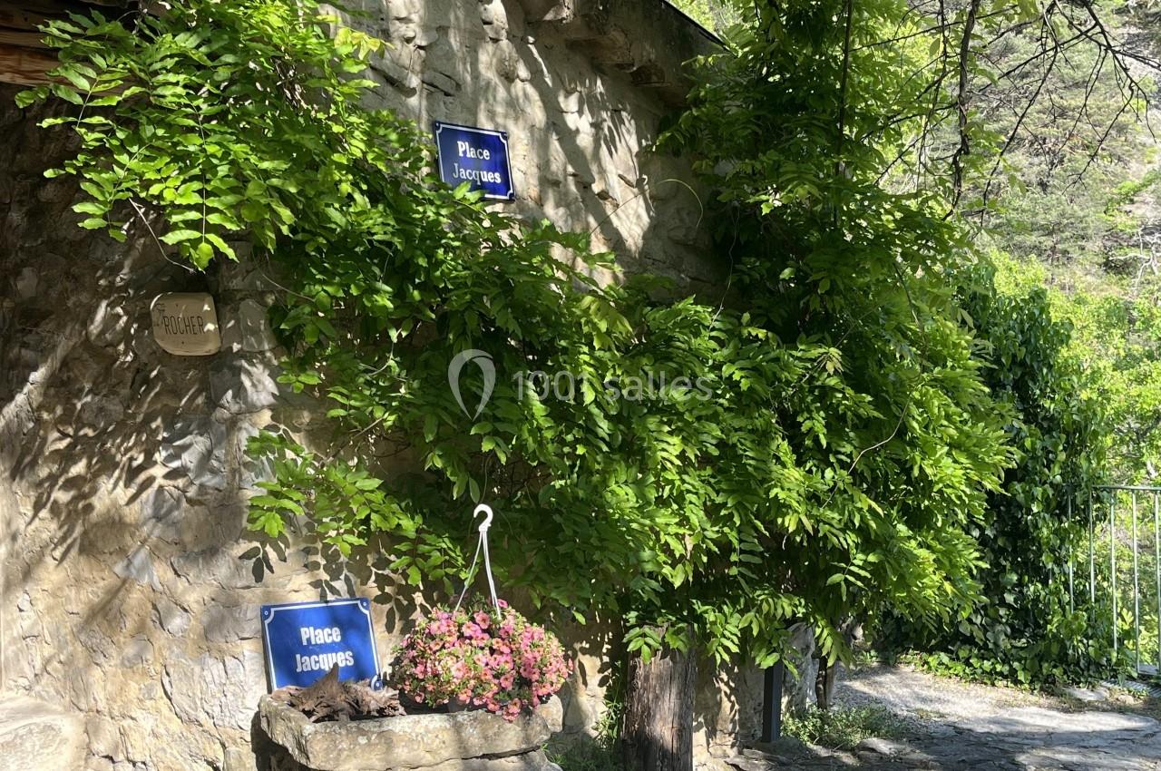 Façade en pierre avec des plantes grimpantes, deux panneaux ’Place Jacques’ et une jardinière fleurie au premier plan.