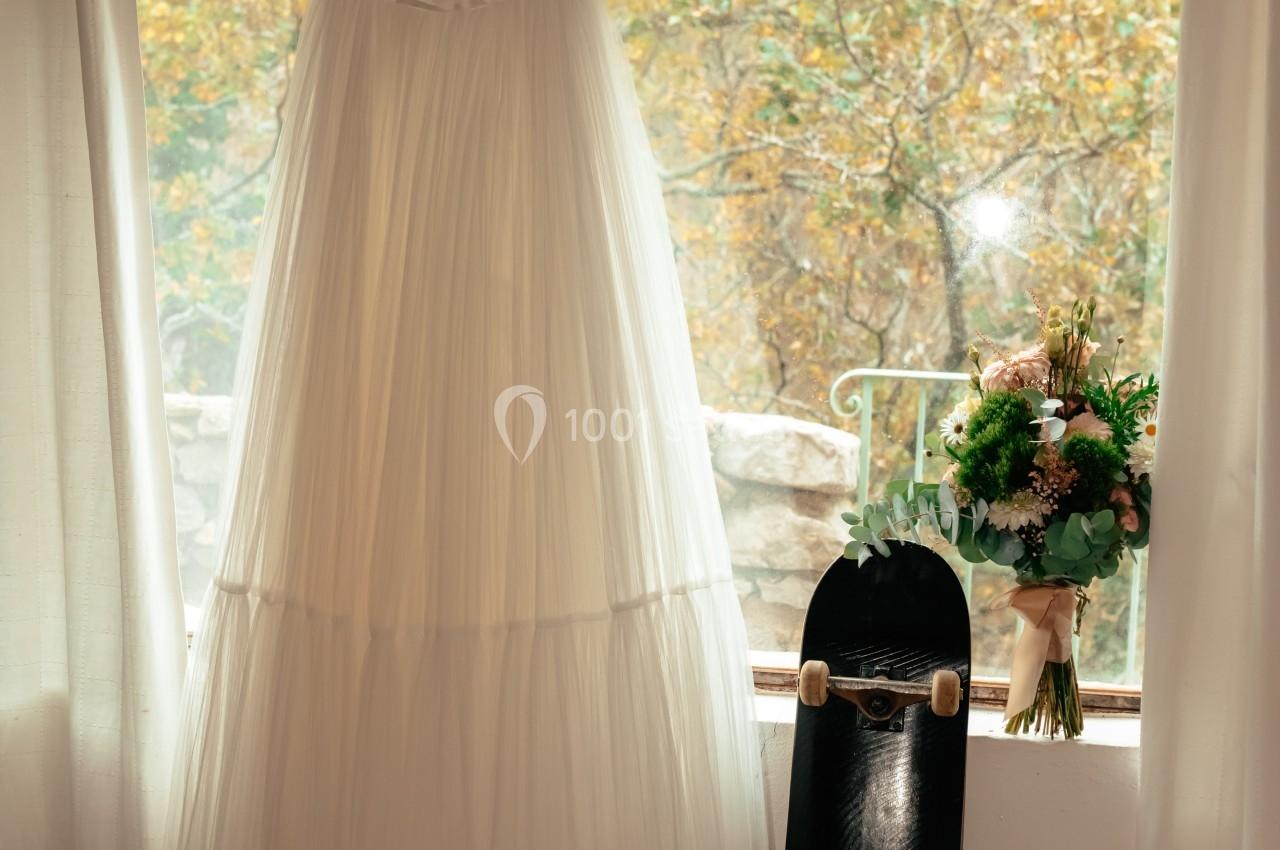Robe de mariée suspendue près d'une fenêtre, avec un bouquet, un skateboard et des chaussures à talons posés au sol.