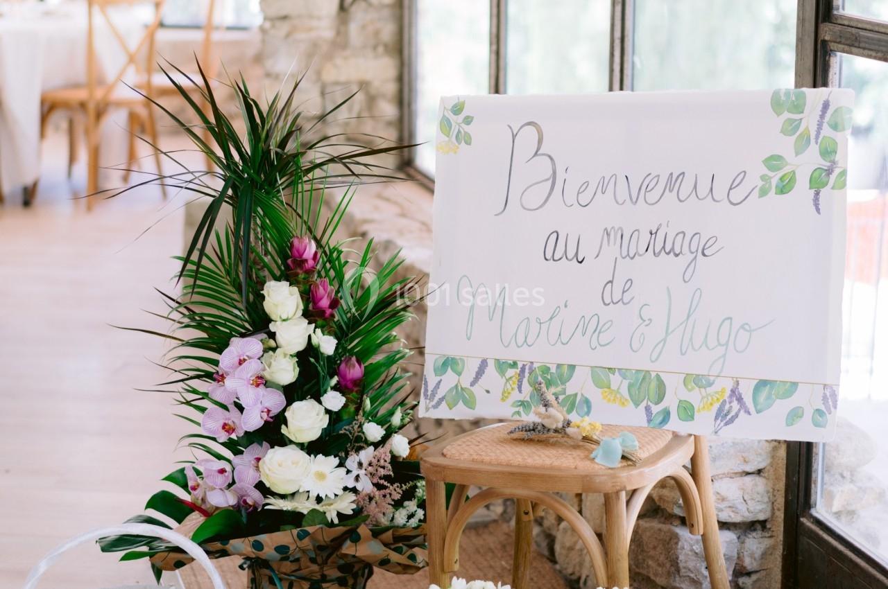 Panneau de bienvenue pour un mariage entouré de fleurs, paniers et plantes dans une salle lumineuse.