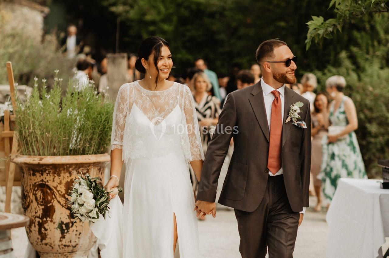 Un couple habillé pour un mariage marche main dans la main dans un jardin extérieur, entouré d'invités.