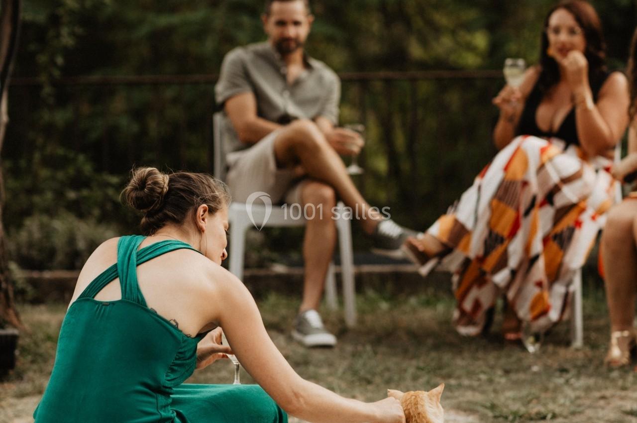 Une femme en robe verte caresse un chat roux, avec des personnes assises en arrière-plan dans un jardin.