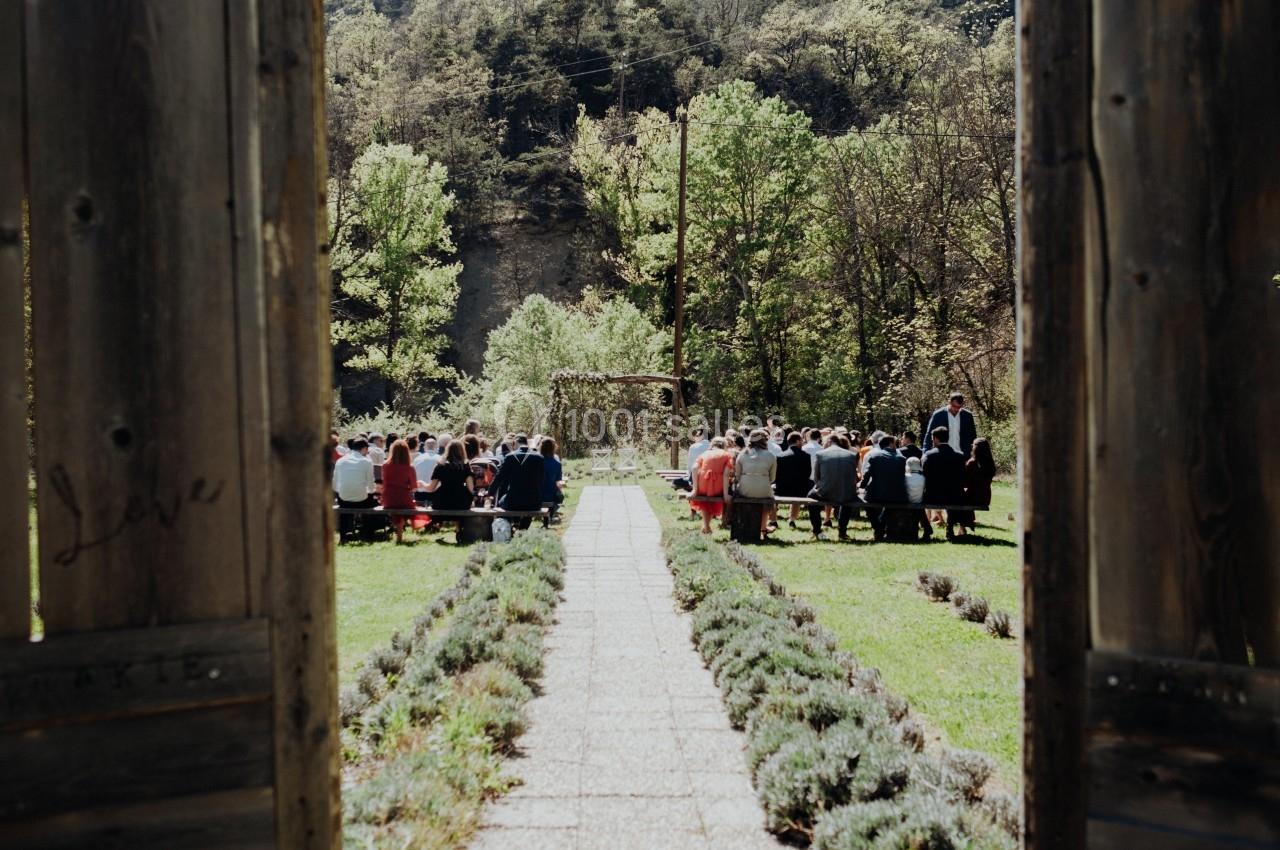 Vue d'une cérémonie en plein air avec des invités assis, entourée de verdure et encadrée par des portes en bois ouvertes.