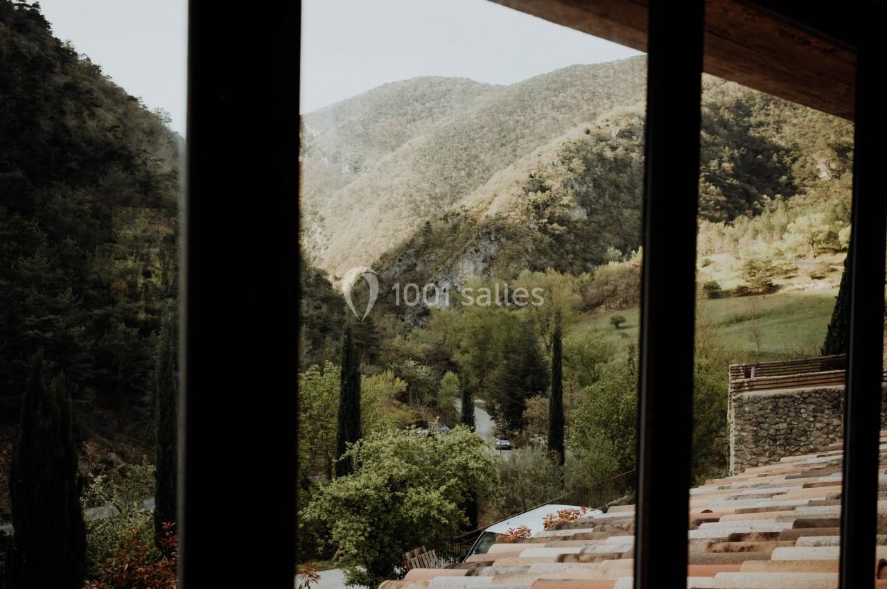 Vue sur des collines verdoyantes et une route bordée d'arbres à travers une fenêtre avec un toit en premier plan.