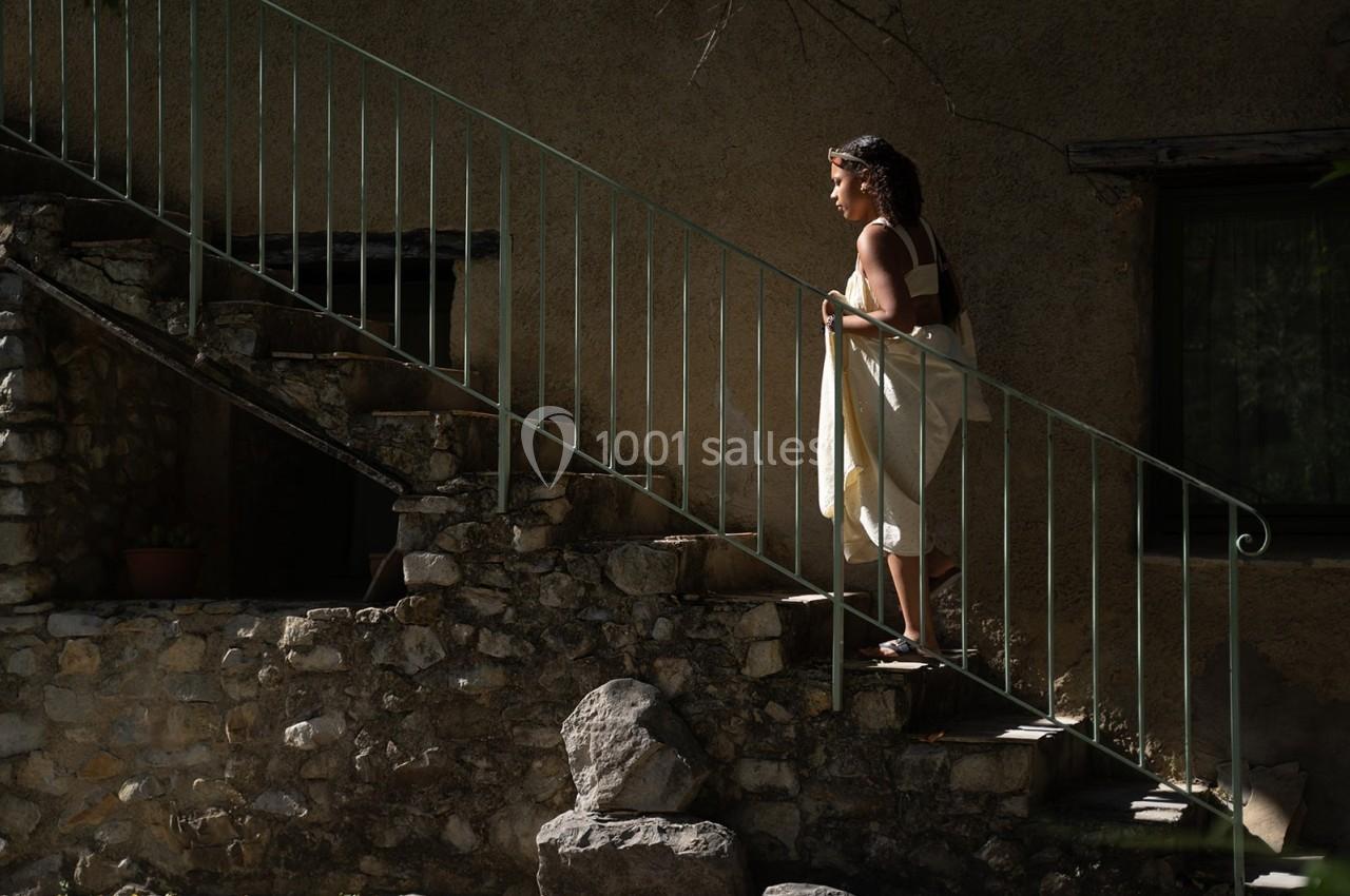 Une femme monte un escalier en pierre à l'extérieur d'une maison, éclairée par une lumière naturelle.