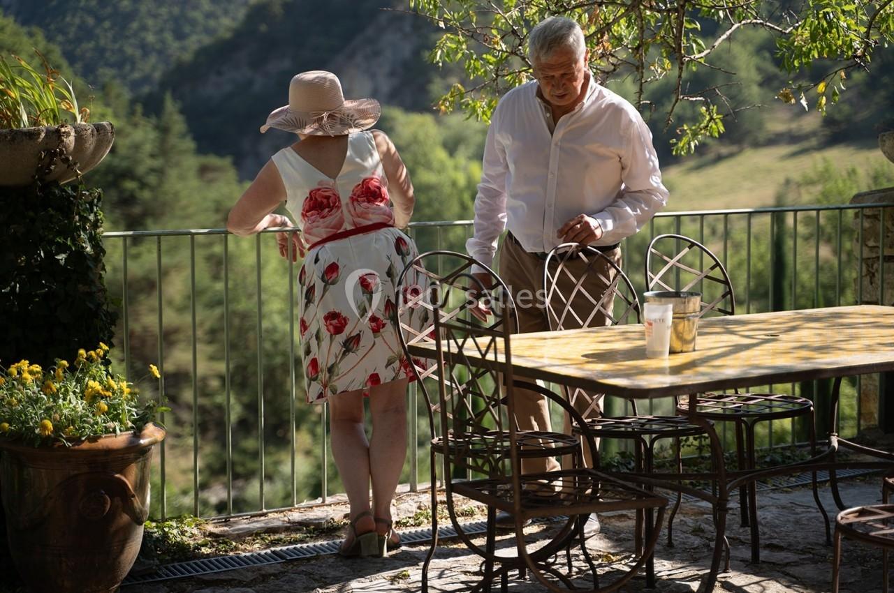 Un homme et une femme discutent sur une terrasse ensoleillée avec vue sur un paysage verdoyant.