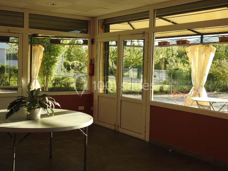 Salle lumineuse avec grandes baies vitrées donnant sur un jardin, table ronde avec plante en pot au premier plan.