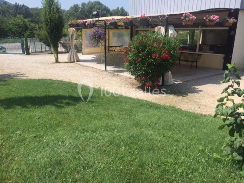 Espace extérieur avec pelouse, terrasse couverte, plantes fleuries et allée gravillonnée près d'un bâtiment.