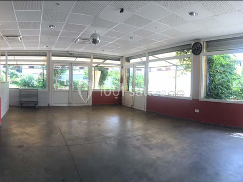 Salle vide avec sol en béton, murs rouges, grandes fenêtres laissant entrer la lumière et vue sur un espace extérieur…