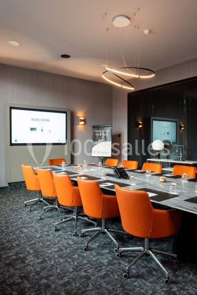 Salle de réunion moderne avec table rectangulaire, chaises orange, écran interactif et éclairage suspendu.