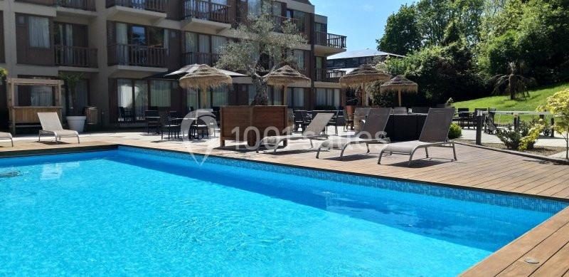 Piscine extérieure entourée de transats et parasols, avec un bâtiment et un jardin en arrière-plan.