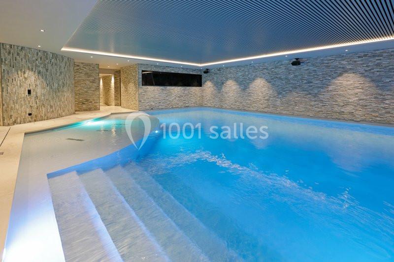 Piscine intérieure moderne avec éclairage tamisé, murs en pierre et plafond à lames métalliques.