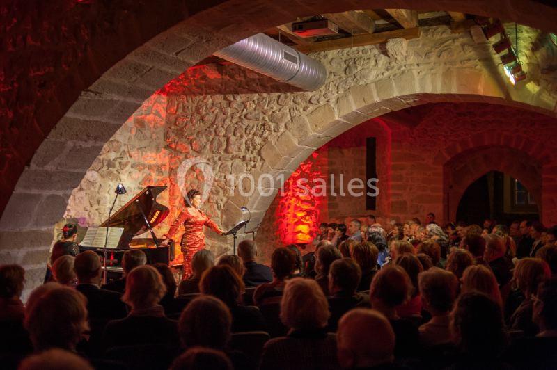 Une chanteuse se produit devant un public dans une salle voûtée en pierre, accompagnée d'un piano.