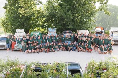 Un grand groupe de personnes en t-shirts verts pose joyeusement devant des véhicules stationnés en extérieur.