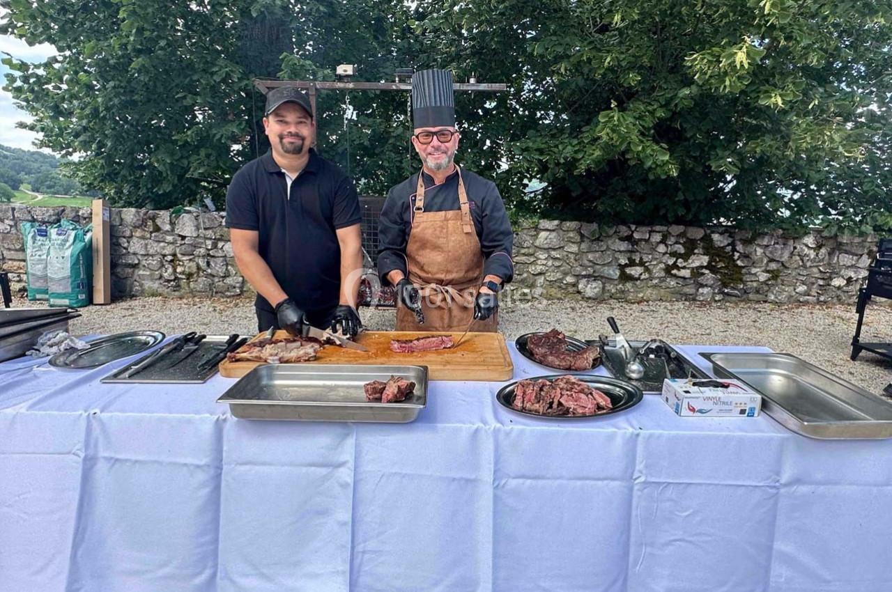 Deux hommes souriants préparent et présentent des morceaux de viande sur une table dressée en extérieur.