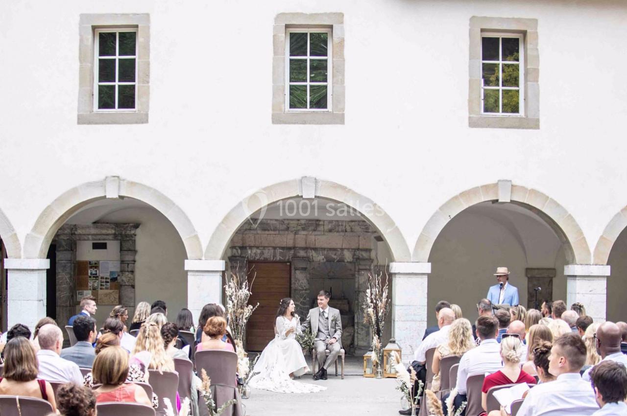 Cérémonie de mariage en extérieur sous des arches, avec les mariés assis face à des invités attentifs.