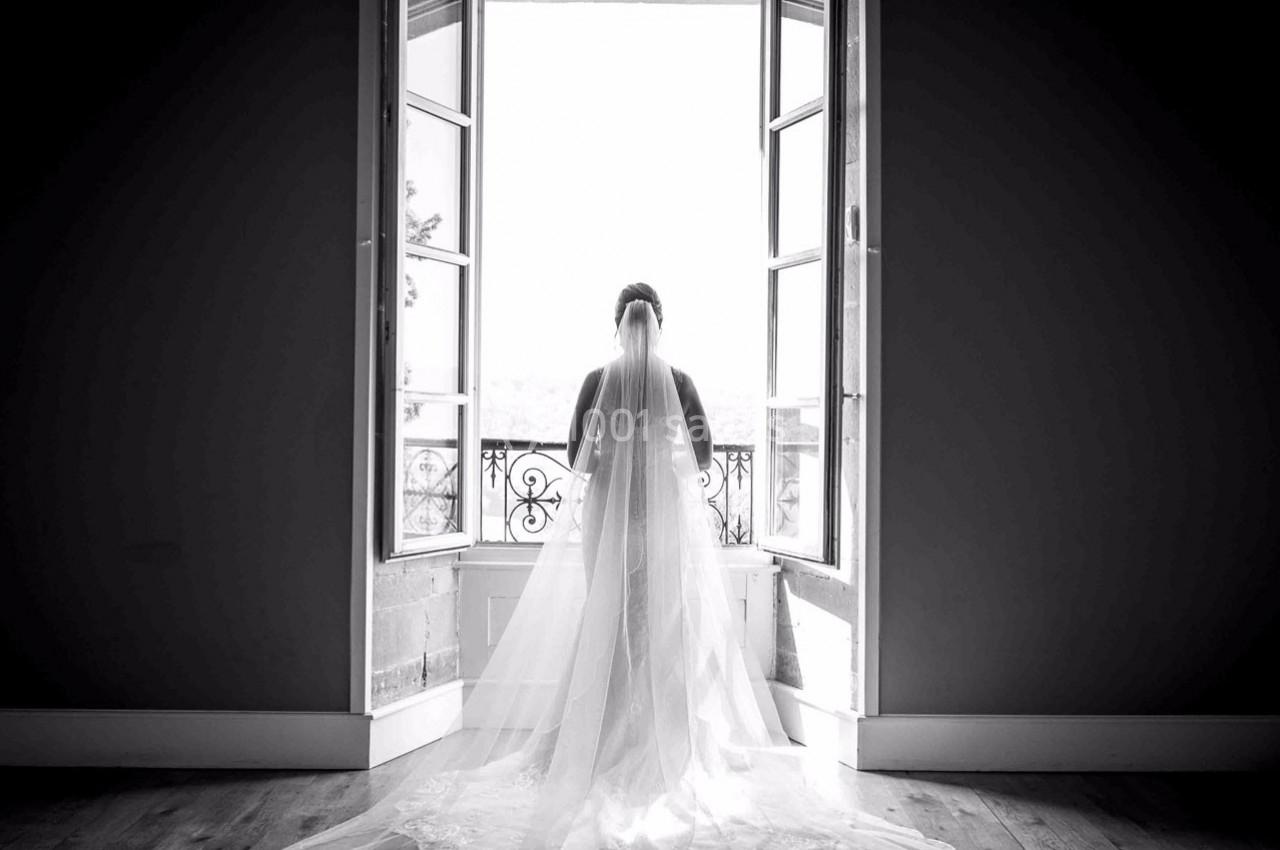 Une mariée en robe blanche et voile regarde par une grande fenêtre ouverte, laissant entrer la lumière naturelle.