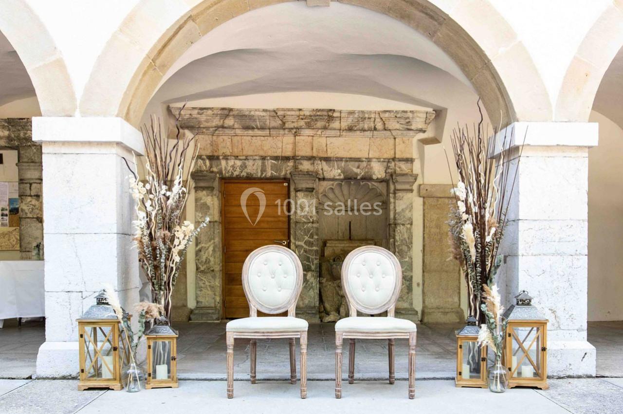 Deux chaises blanches à dossier capitonné devant une porte en bois, entourées de lanternes et de décorations florales.