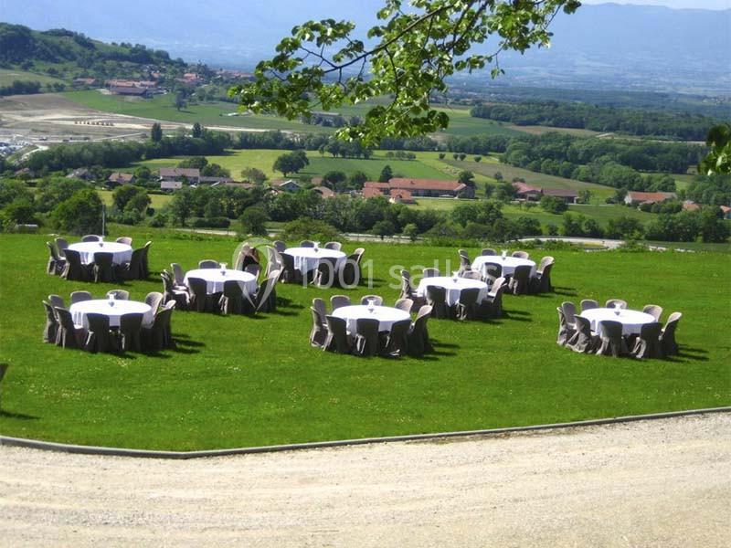 Tables rondes dressées avec nappes blanches et chaises couvertes, disposées sur une pelouse avec vue sur la campagne.