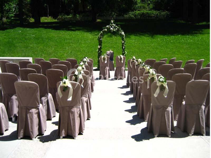Chaises alignées avec housses beiges et arche fleurie au fond, installées en extérieur sur une pelouse pour une cérémonie.