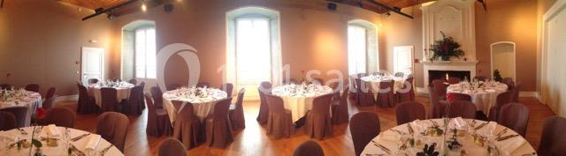 Salle de réception lumineuse avec tables rondes dressées, chaises couvertes et cheminée décorée de fleurs.