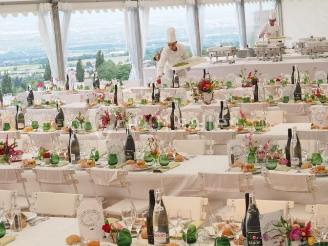 Tables dressées pour un événement avec nappes blanches, décor floral, bouteilles de vin et un chef en action.