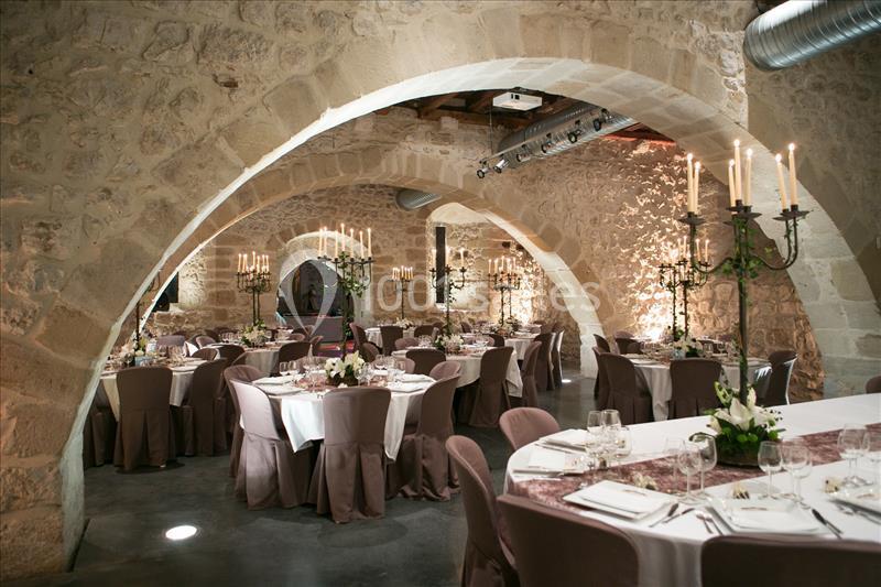 Salle voûtée en pierre avec tables rondes dressées pour un événement, éclairée par des chandeliers.