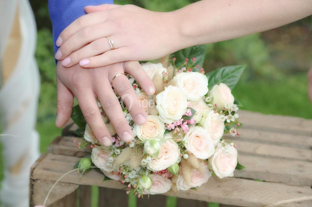 Deux mains avec des alliances posées sur un bouquet de roses blanches et roses, sur une table en bois.