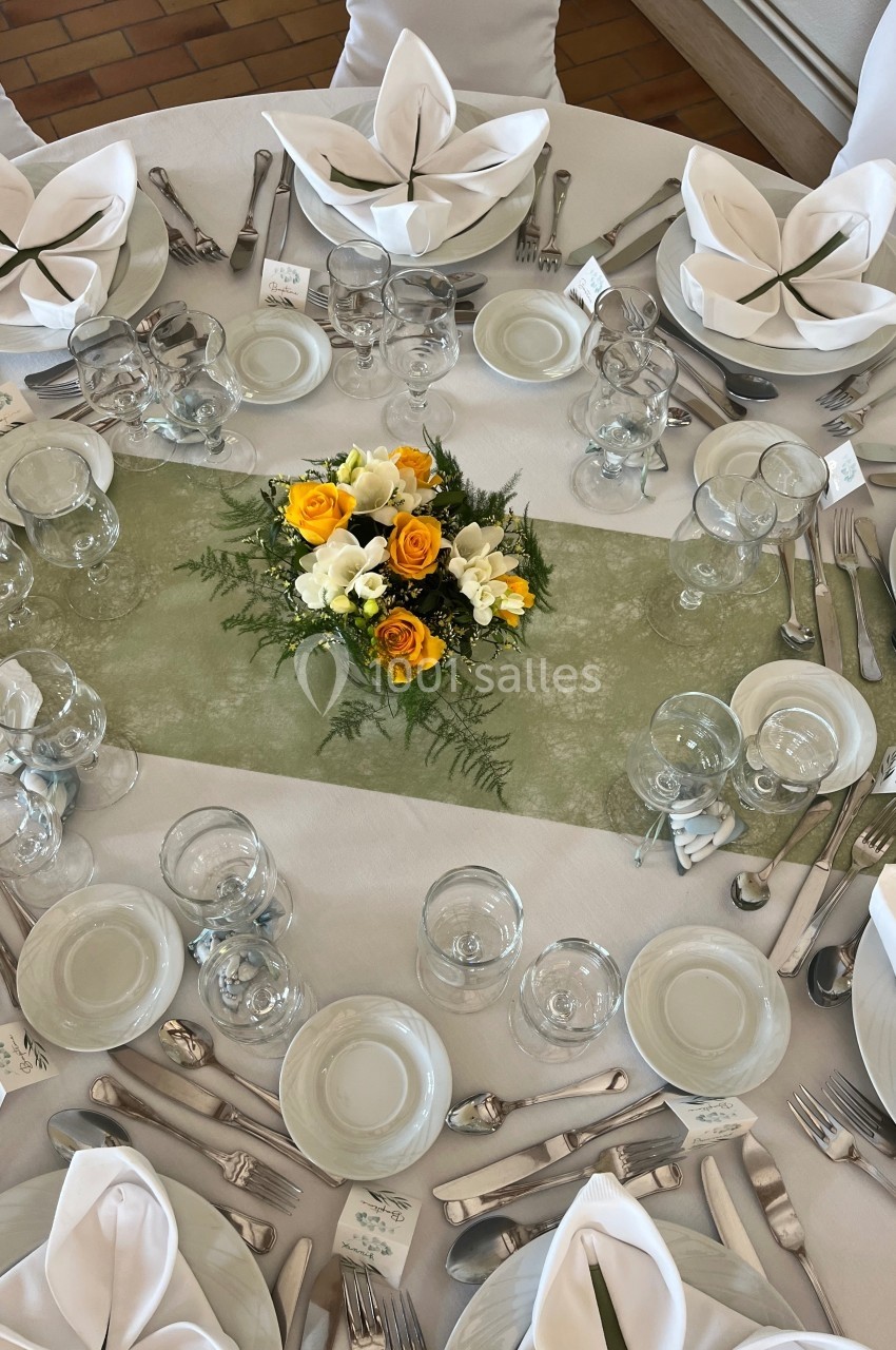Table dressée avec nappes blanches, serviettes pliées, couverts alignés et centre de table floral jaune et blanc.
