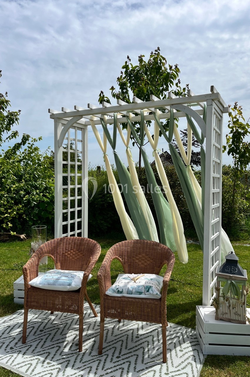 Deux fauteuils en rotin sous une pergola blanche décorée de voiles pastel, installés sur une pelouse avec un tapis.