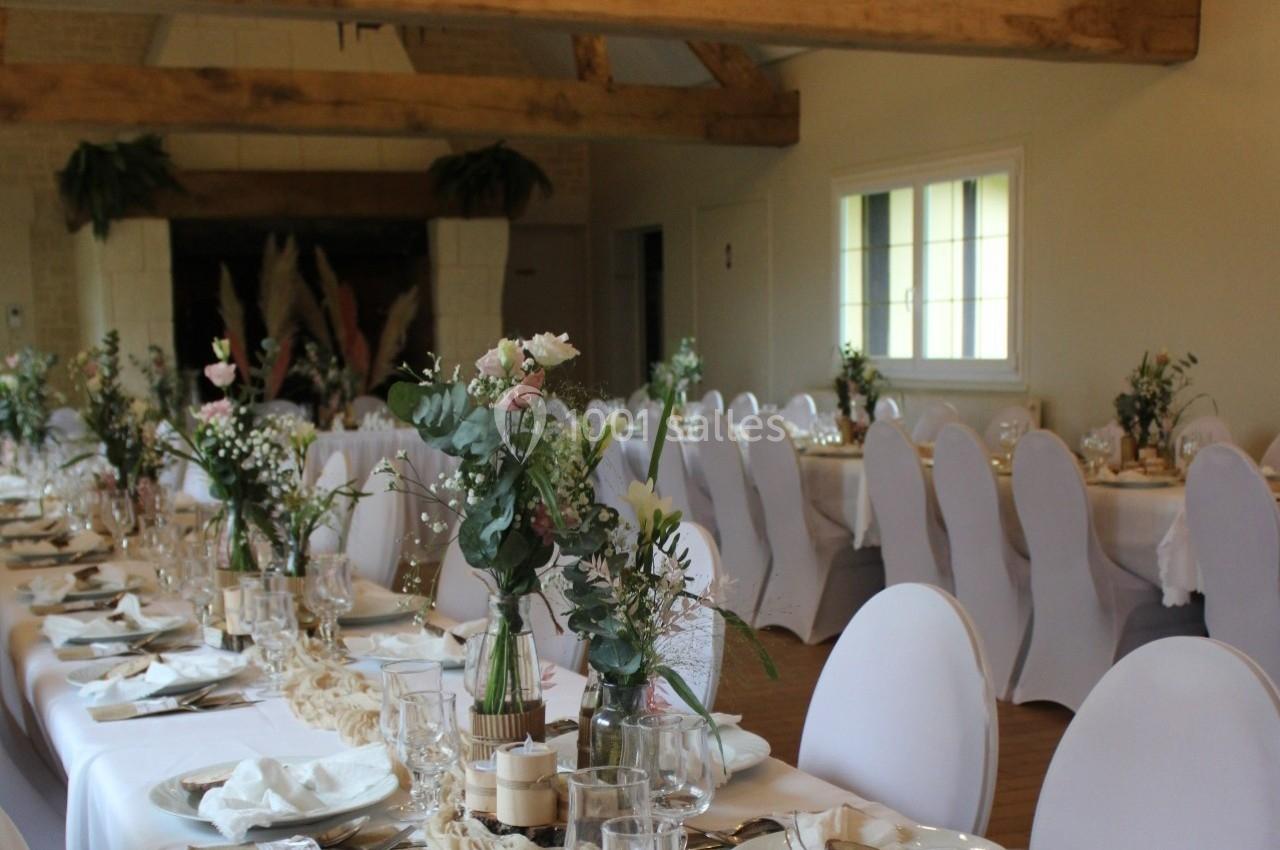 Salle de réception décorée avec des tables alignées, nappes blanches, chaises houssées et centres de table fleuris.
