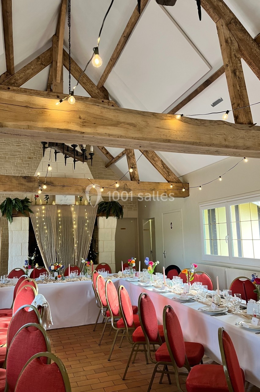 Salle de réception décorée avec des tables dressées, chaises rouges, poutres apparentes et guirlandes lumineuses.
