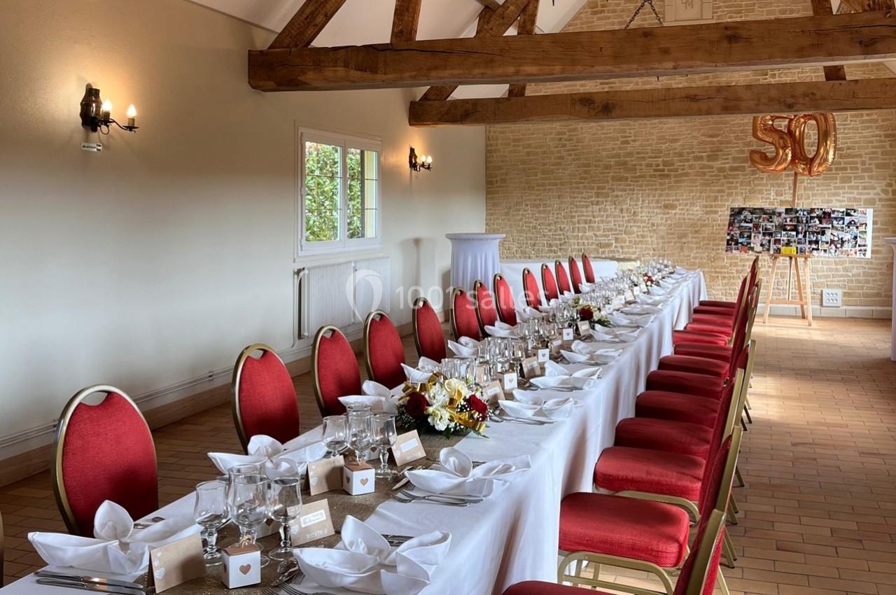 Salle décorée pour une réception avec une longue table dressée, chaises rouges et ballons dorés formant ’50’.