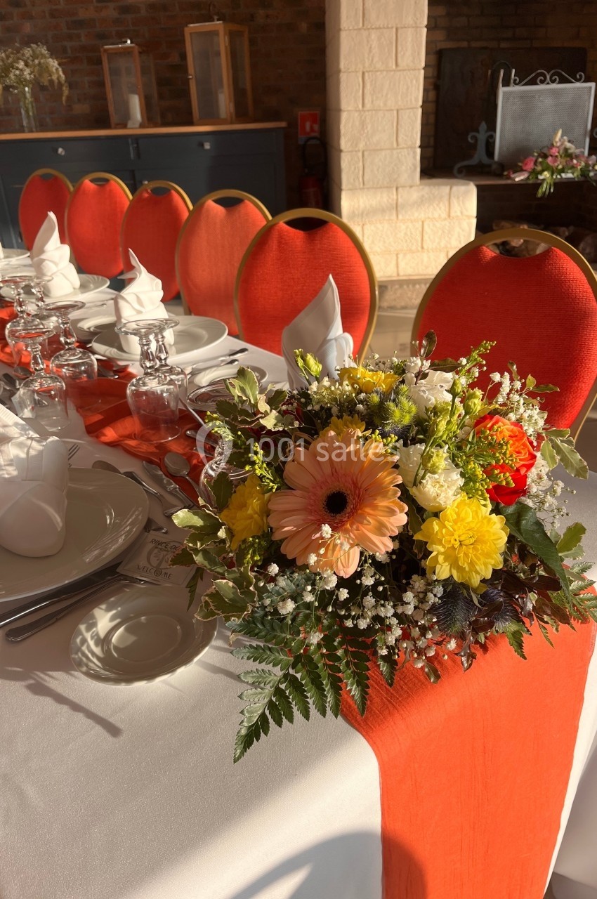 Table dressée avec nappes blanches, chemin de table orange, vaisselle élégante et bouquet de fleurs colorées au centre.