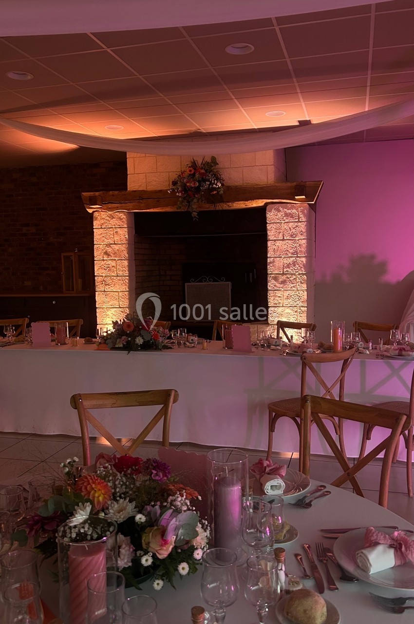 Salle de réception décorée avec des tables dressées, des fleurs et un éclairage chaleureux près d'une cheminée en pierre.