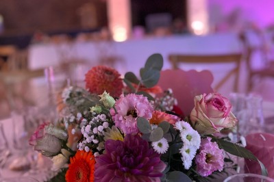 Table ronde élégamment dressée avec vaisselle, couverts, serviettes roses et centre de table floral coloré.