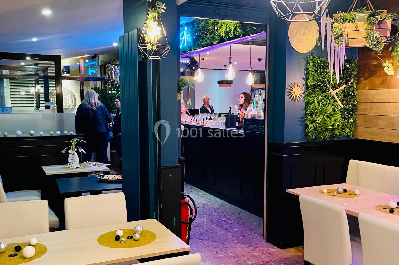 Intérieur d'un restaurant moderne avec tables dressées, décoration végétale et vue sur un bar animé en arrière-plan.