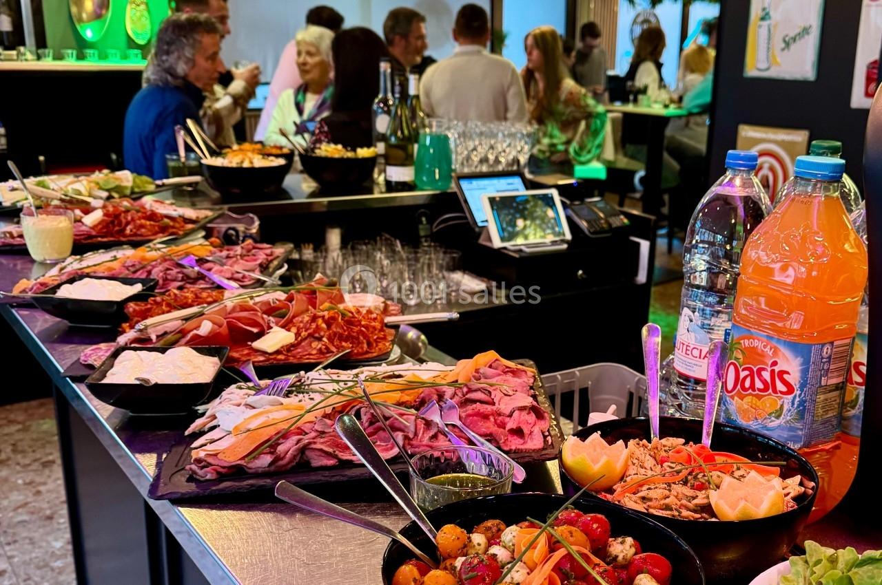 Buffet varié avec charcuterie, légumes marinés et boissons, dans un bar animé avec des convives en arrière-plan.