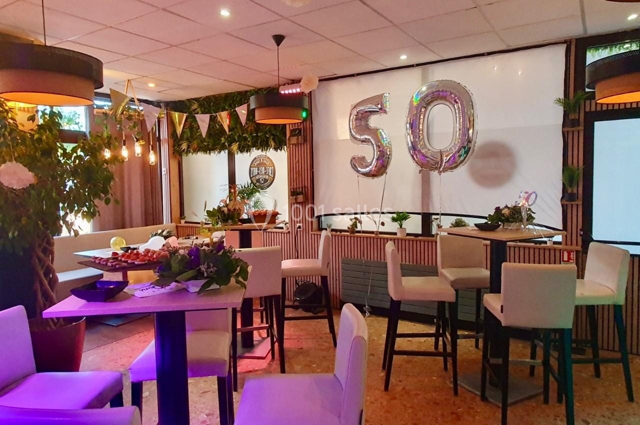 Salle décorée pour une fête avec des ballons ’50’, des guirlandes, des plantes et des tables hautes entourées de chaises.