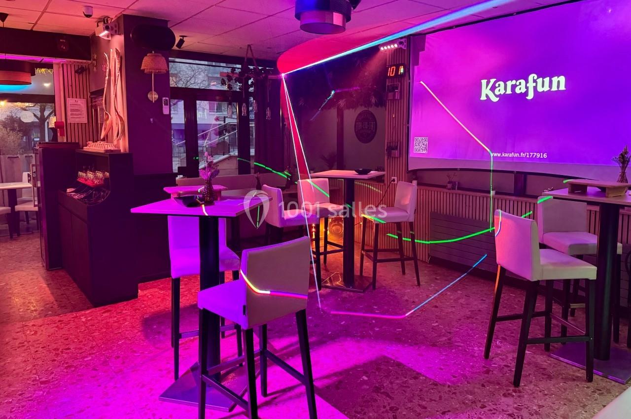 Salle de karaoké avec tables hautes, chaises blanches, éclairage coloré et écran affichant ’Karafun’.